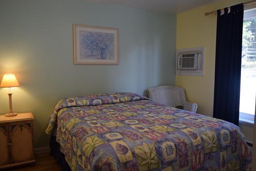 A wide view of a guest room with a bed, nightstand, lamp, window, and air conditioner.